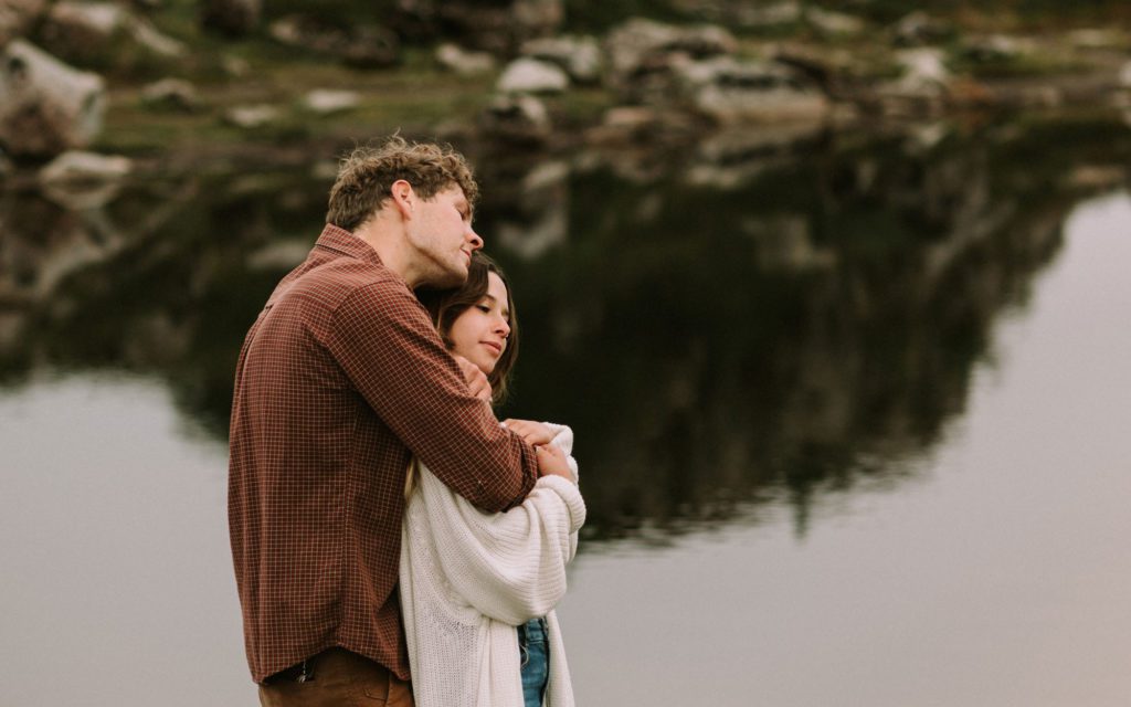 A couple stands in front of an alpine lake reflection at a photoshoot.