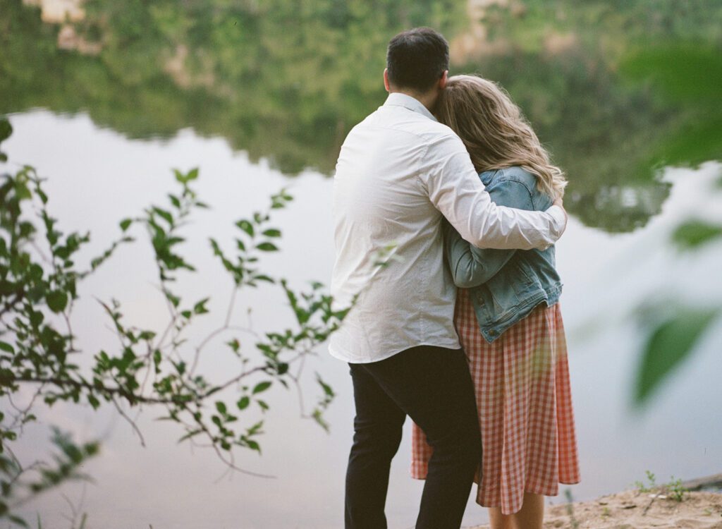 Couple cuddles by the river at neighborhood engagement session.